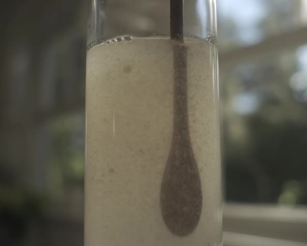 A spoon stirs cloudy particles suspended in liquid within a glass vial in a close-up shot