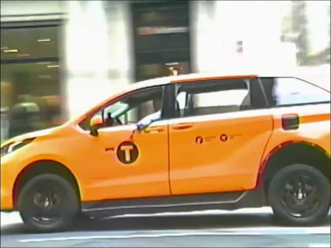 A yellow taxi cab drives through a city intersection as pedestrians cross in grainy, retro-style footage