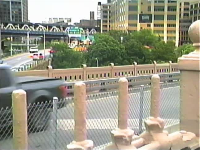 A slow zoom out from a green highway sign for the Brooklyn Bridge exit reveals a bustling, multi-level roadway in New York City