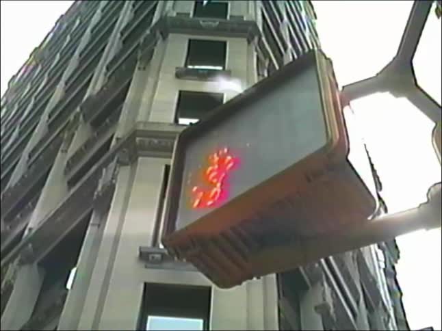 A low-angle shot shows a pedestrian crossing signal changing from a red 'don't walk' hand to a white 'walk' person symbol on a city street