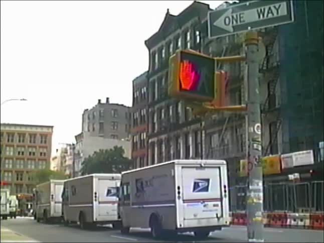 Vintage footage shows a line of USPS mail trucks parked on a city street as a pedestrian light cycles