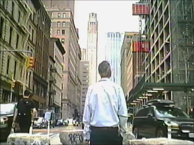 A man stands in the middle of a New York City street, looking up at the towering skyscrapers in vintage VHS footage