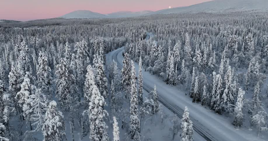 Aerial footage follows a lone car driving down a winding road through a vast, snow-covered forest at sunset