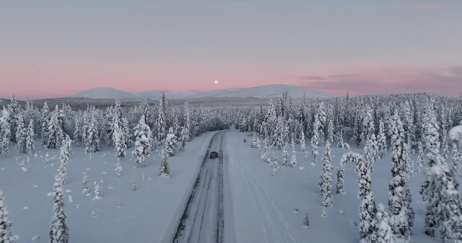 Aerial shot of a car driving on a remote snowy road through a winter wonderland forest at dusk, with a beautiful pink sky and full moon