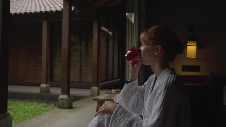 A serene woman in a white robe sips from a red cup while looking out at a peaceful courtyard from her dimly lit hotel room