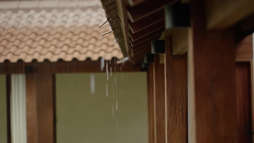 Raindrops fall in slow motion from the eaves of a traditional wooden building with a tiled roof