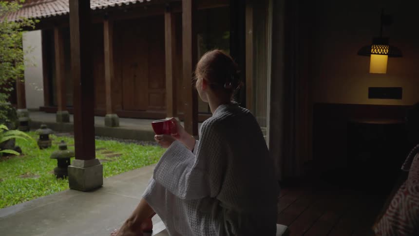 A serene woman in a white bathrobe enjoys a warm beverage while sitting by an open door overlooking a tranquil traditional Asian courtyard