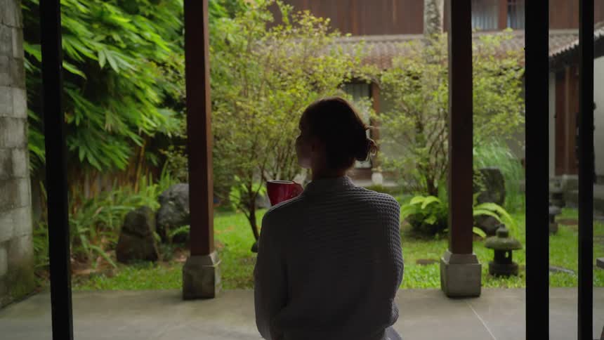 A woman in a bathrobe peacefully sips her morning coffee while sitting in a doorway, enjoying the view of a serene, rainy Asian garden
