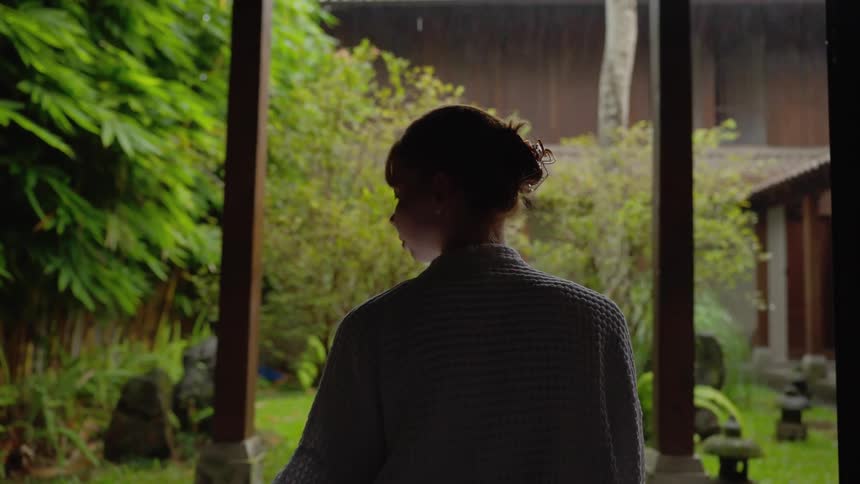 A woman in a bathrobe stands on a tranquil patio, drinking tea and watching the rain fall in a lush tropical garden