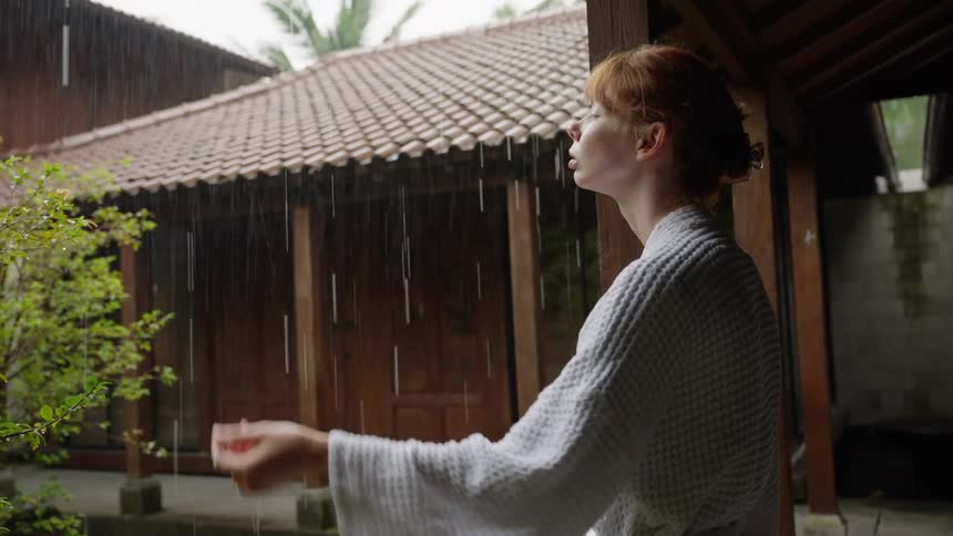 A serene woman in a white bathrobe reaches out her hand to feel the rain at a tranquil spa resort