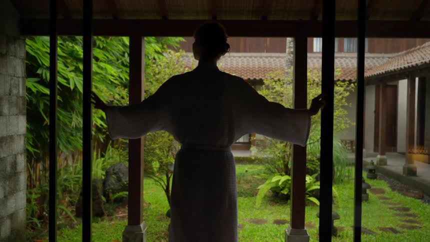 A woman in a white bathrobe slowly opens the doors of her room to reveal a beautiful and serene Asian-style garden during a light rain
