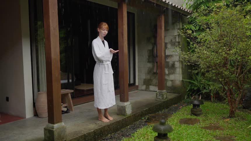 A young woman in a white bathrobe stands on the porch of her tropical villa, peacefully watching the rain fall in the lush garden