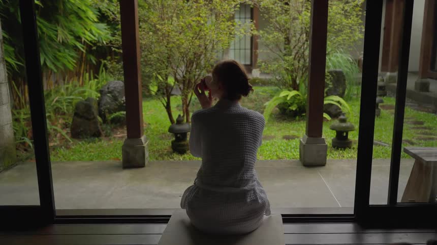 A woman in a white bathrobe enjoys a quiet moment with a cup of tea while gazing out at a tranquil green garden