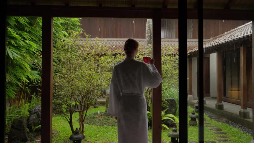 Woman in a white bathrobe enjoys a warm drink on her villa patio while watching the rain fall in a lush, peaceful garden