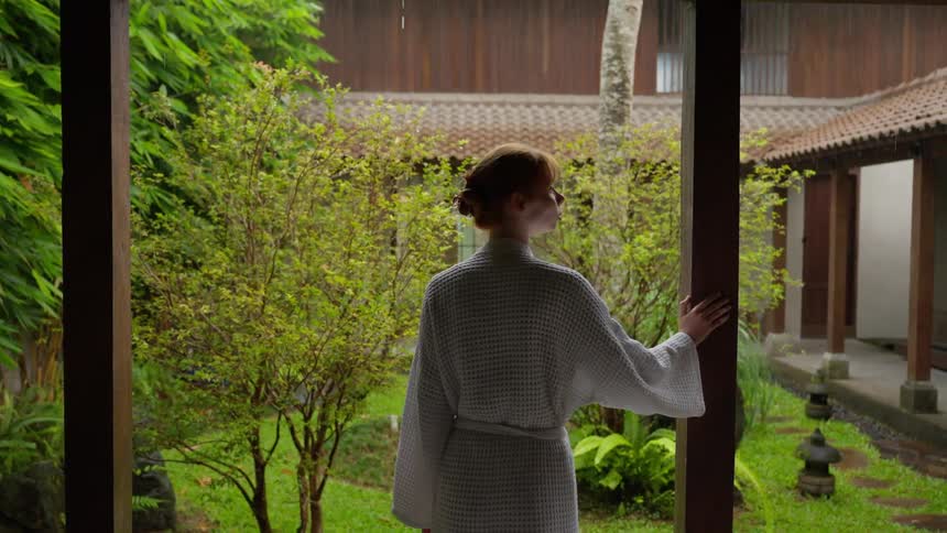 A woman in a white bathrobe stands on a covered patio, peacefully watching the rain fall in a lush Asian-style courtyard