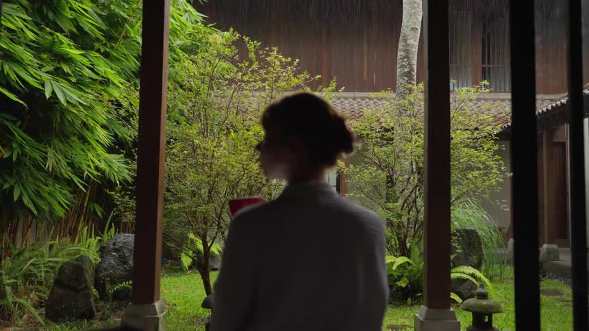 From behind, a woman in a robe enjoys a hot drink while watching the rain fall in a tranquil, lush garden