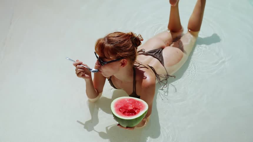 High angle shot of a woman in a bikini relaxing in a pool and eating a watermelon in slow motion