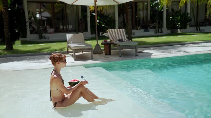 A young woman in a bikini relaxes in the shallow end of a beautiful turquoise swimming pool holding a watermelon