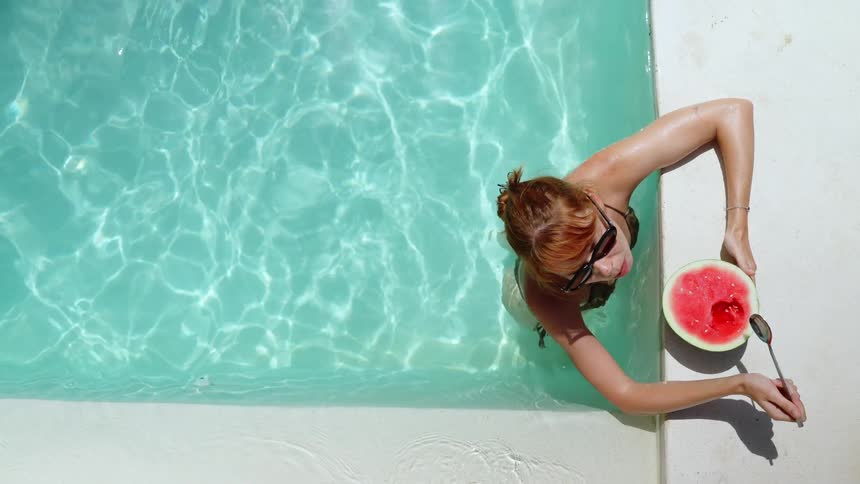 Top-down view of a redhead woman in a bikini relaxing in a sunlit swimming pool while eating a fresh watermelon