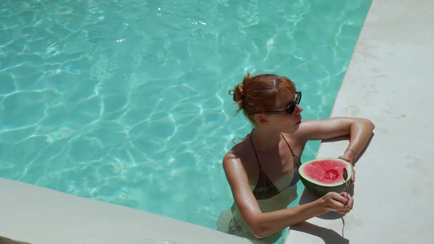 A woman with red hair and sunglasses relaxes in a turquoise swimming pool while holding a watermelon and a spoon