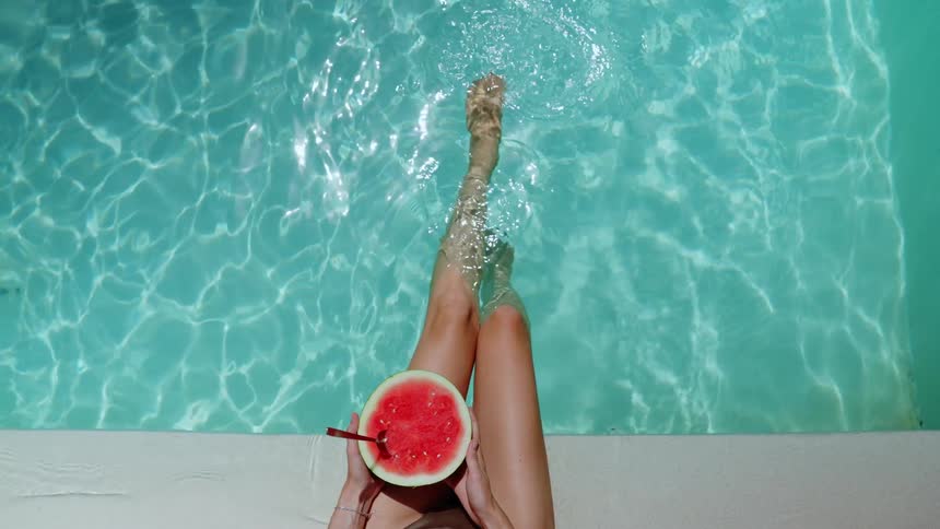 Top-down point of view of a woman relaxing in a turquoise pool with a fresh watermelon smoothie