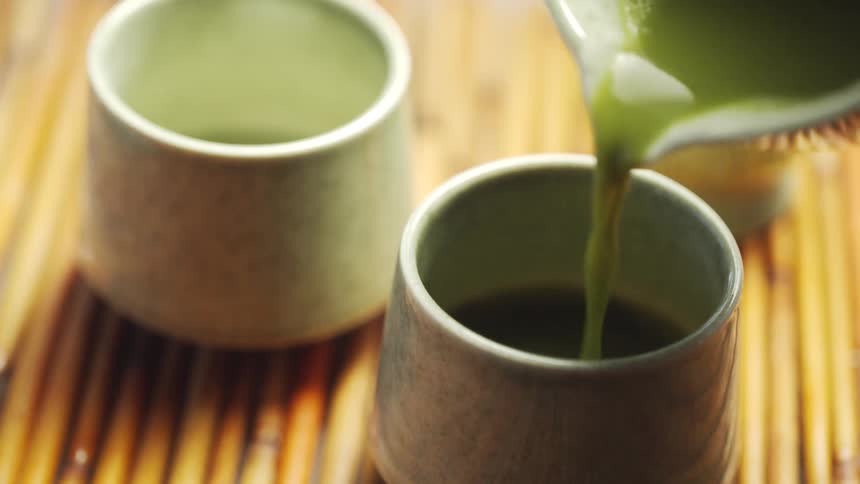 A close-up slow-motion shot of pouring vibrant matcha green tea into a Japanese ceramic teacup on a bamboo mat
