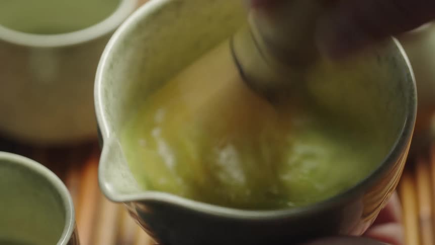 Close up of a hand whisking matcha powder with a traditional bamboo whisk in a ceramic bowl to make Japanese green tea
