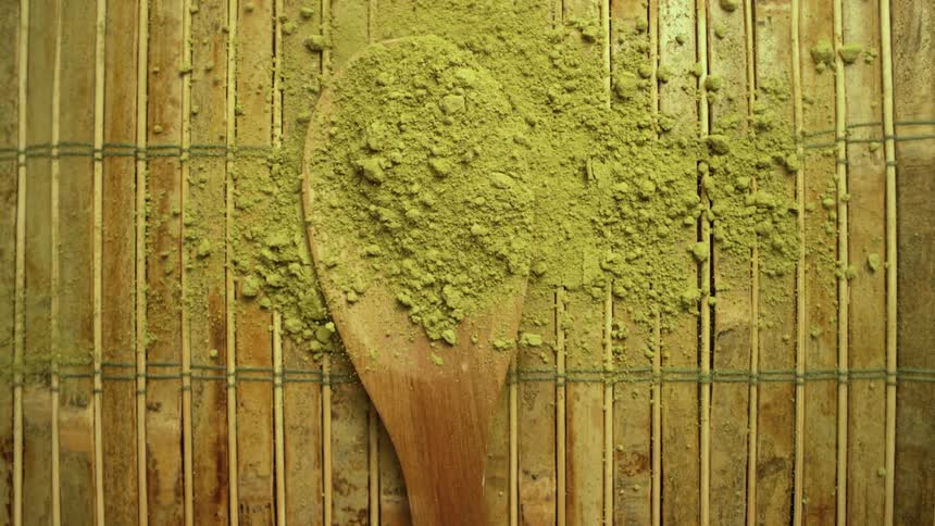 Green matcha powder falls in slow motion onto a wooden spoon against a rustic bamboo background