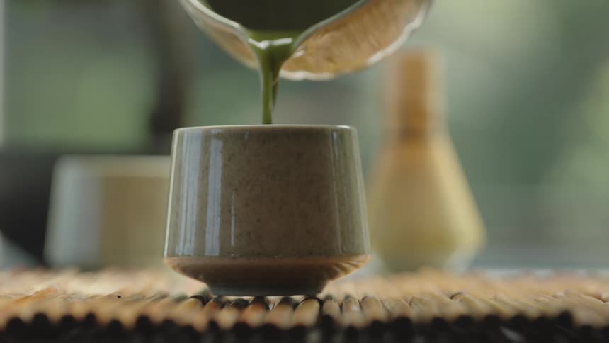 Close up shot of Japanese matcha tea being slowly poured into a ceramic cup on a bamboo mat