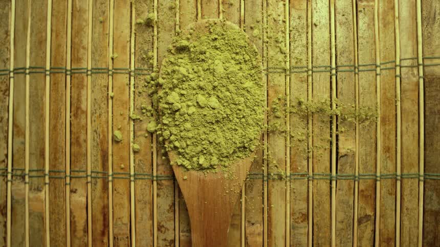 Vibrant green matcha powder gently falls onto a wooden spoon in a slow-motion, top-down view over a bamboo mat