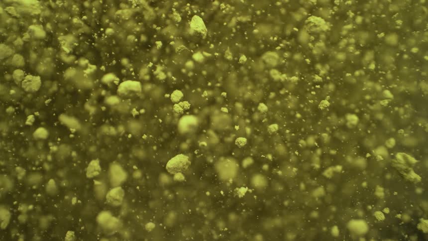 Macro shot of fine yellow-green powder particles slowly floating and dispersing in a liquid against a dark background