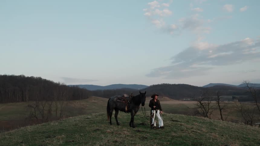 A cowgirl stands with her loyal horse on a scenic grassy hill, overlooking a vast valley at dusk