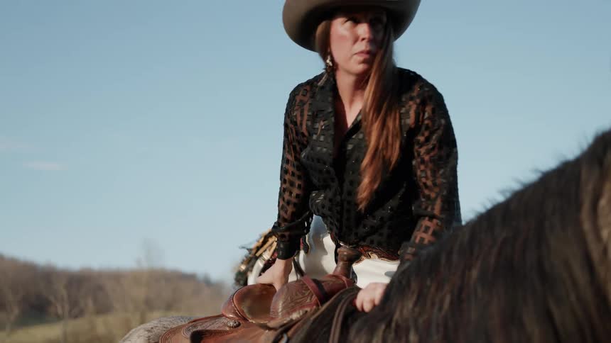 A cowgirl in a western-style outfit slowly mounts her horse on a sunny day in the countryside