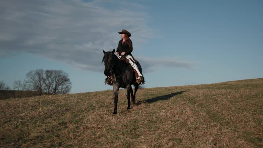 A woman in a cowboy hat gracefully rides her majestic black horse up a grassy hill under a beautiful blue sky