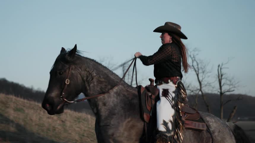A cowgirl in a cowboy hat and decorative chaps rides her powerful gray horse across a grassy hill in slow motion