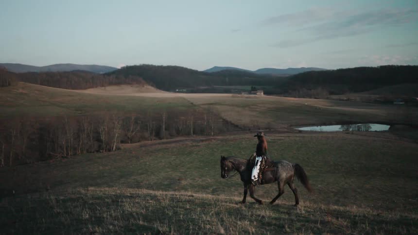 A lone cowgirl rides her horse in slow motion across a grassy hilltop at dusk, overlooking a scenic valley with rolling hills and a pond