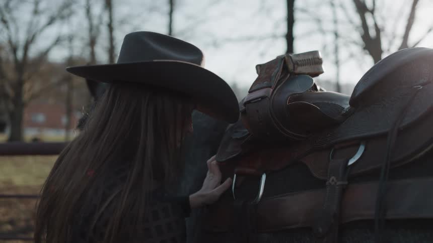 A long-haired woman in a cowboy hat carefully adjusts the leather saddle on her horse outdoors in the afternoon sun