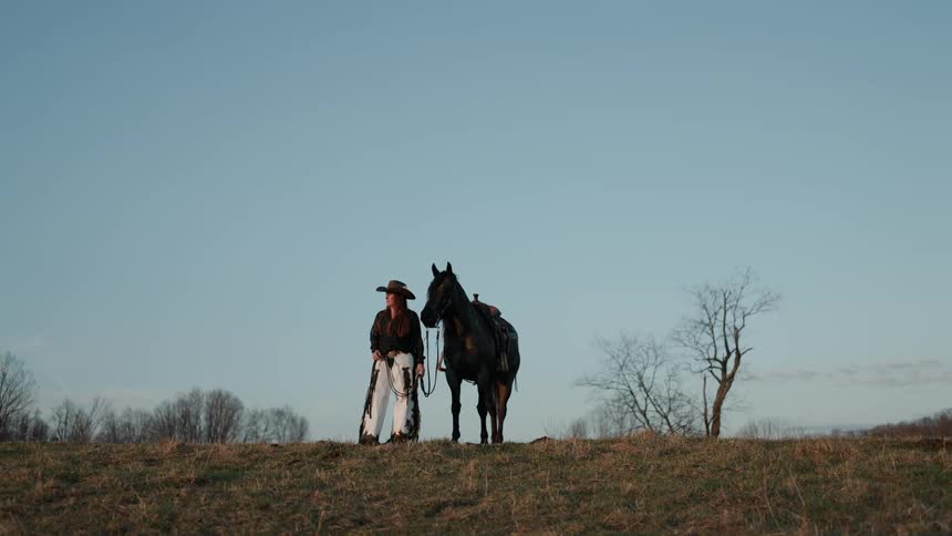 A classic wide shot of a cowgirl and her horse standing together on a rural hilltop at sunset