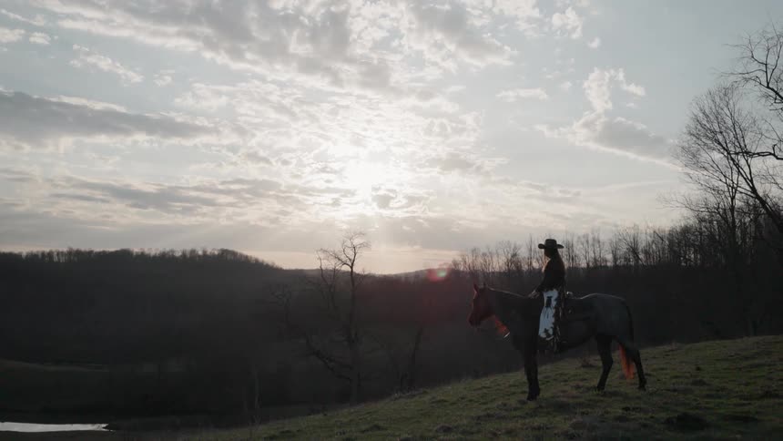 Silhouetted cowgirl on her horse stands on a grassy hill, gazing at the beautiful sunset over the mountains