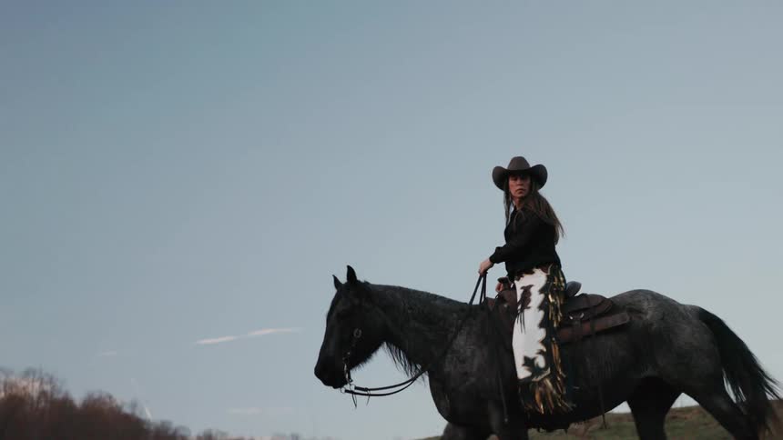 A stylish cowgirl wearing a hat and fringed chaps confidently rides her powerful grey horse up a grassy hill in a majestic slow-motion shot