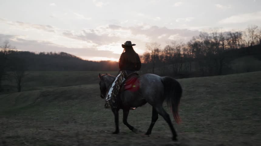 A lone cowgirl on a roan horse slowly rides across a field of rolling hills towards the setting sun