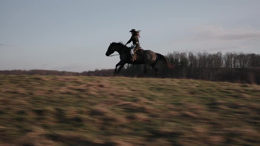 A cowgirl with long flowing hair rides a powerful horse at a fast gallop through a rural field in cinematic slow motion