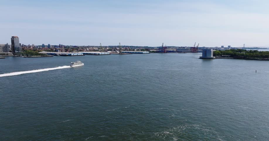 A static aerial view shows a white ferry boat cruising down the river past the industrial Brooklyn shoreline and port on a bright, sunny day