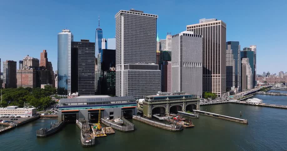 A stunning aerial shot pulls back from the Staten Island Ferry terminal, revealing the spectacular Lower Manhattan skyline and waterfront on a sunny day