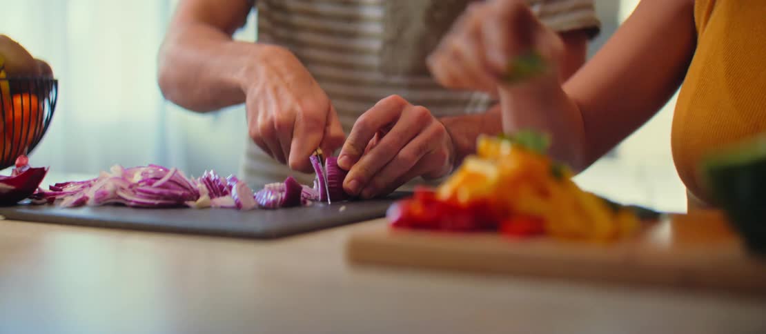 Close-up shot of a couple's hands preparing a meal together, dicing a red onion and colorful peppers on cutting boards