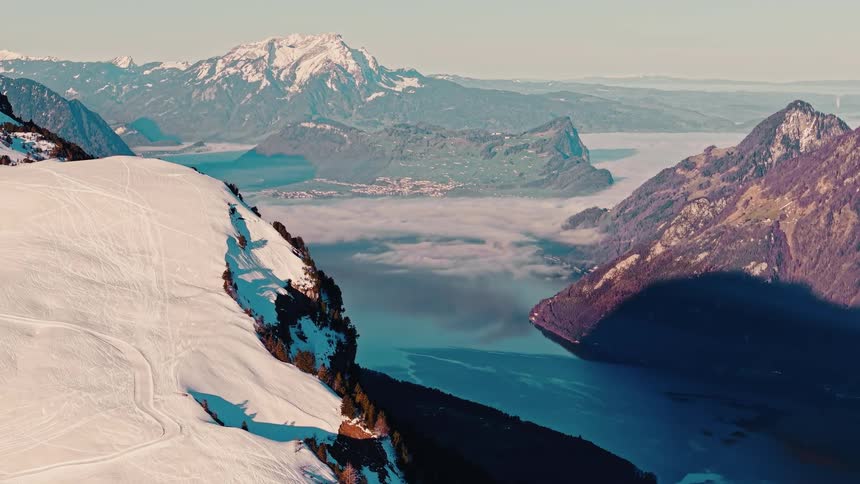 A stunning aerial view from a snowy mountain ridge overlooks a tranquil blue lake and cloud-filled valley in the Swiss Alps