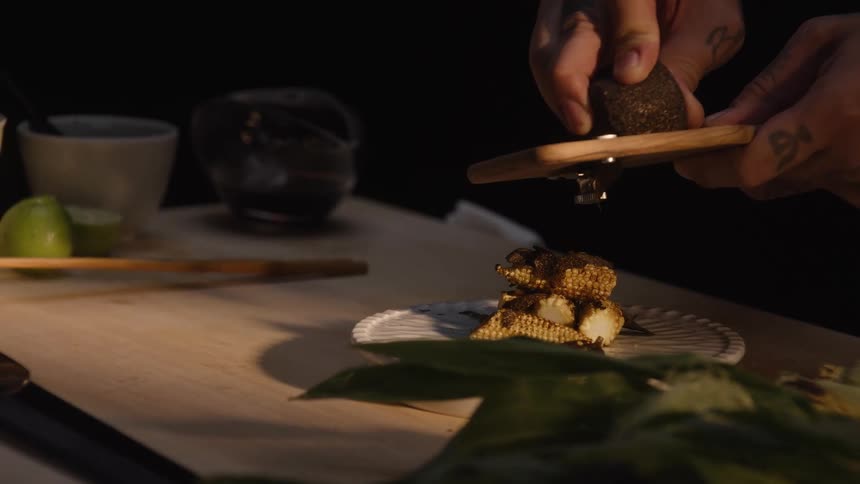 A tattooed chef in slow motion carefully shaves fresh black truffle onto a gourmet dish of baby corn
