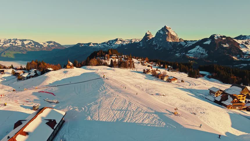 An aerial drone slowly flies over a charming ski village in the Swiss Alps during a beautiful golden hour sunrise