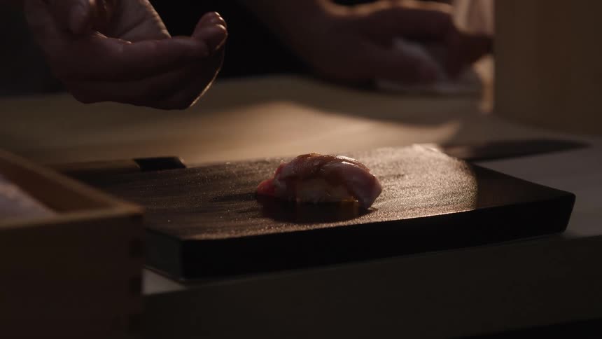 In a dimly lit restaurant, a skilled chef meticulously places a freshly made piece of tuna nigiri on a serving block