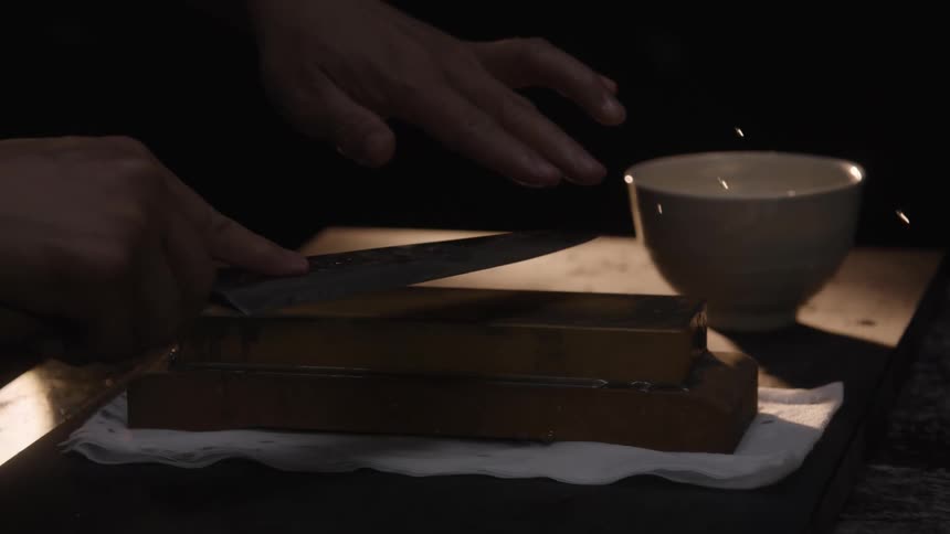 A person pours water over a whetstone and begins sharpening a knife blade in dramatic, moody slow motion